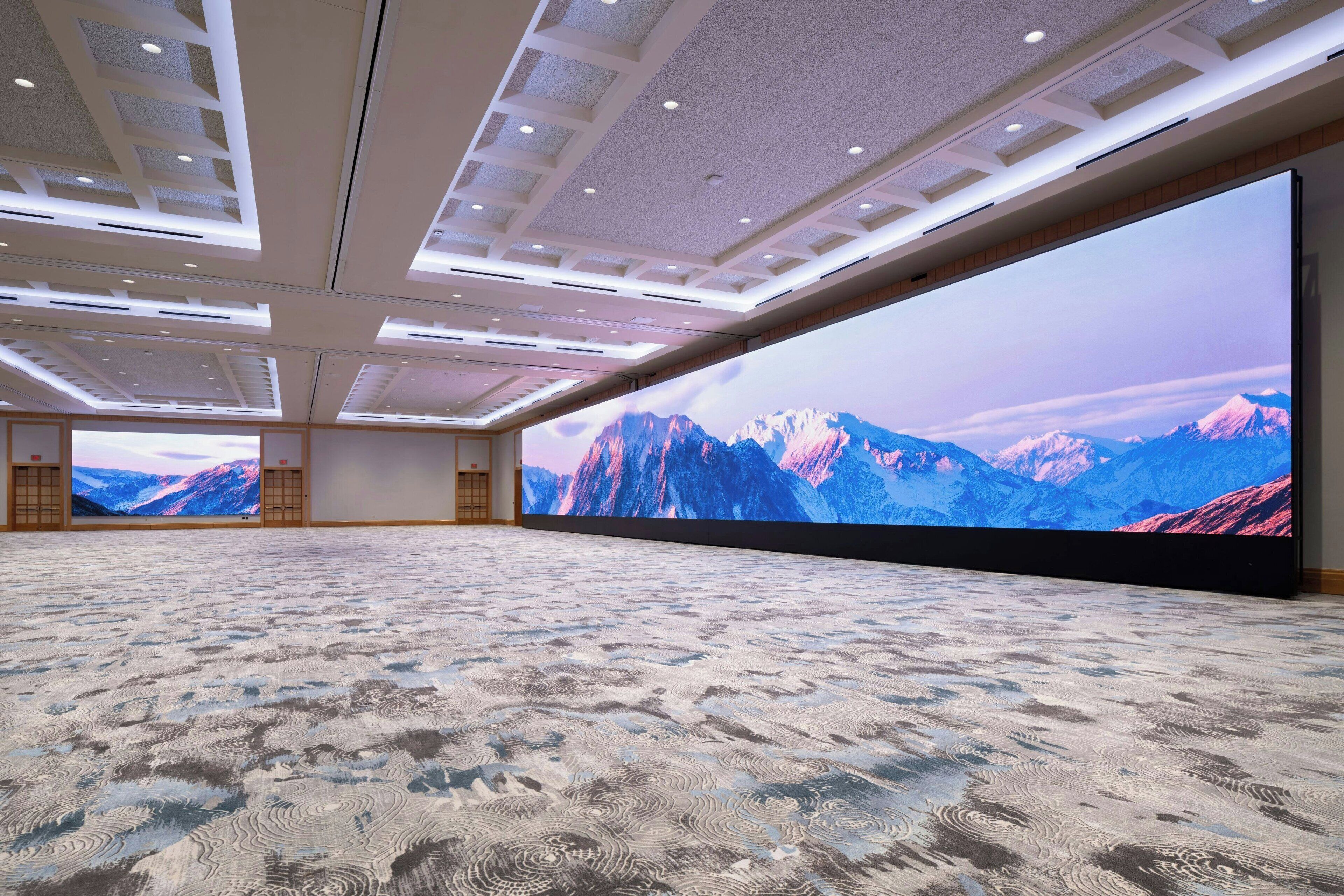 A spacious room with a large carpeted floor, a high ceiling with recessed lighting, and a large screen displaying a mountain landscape.