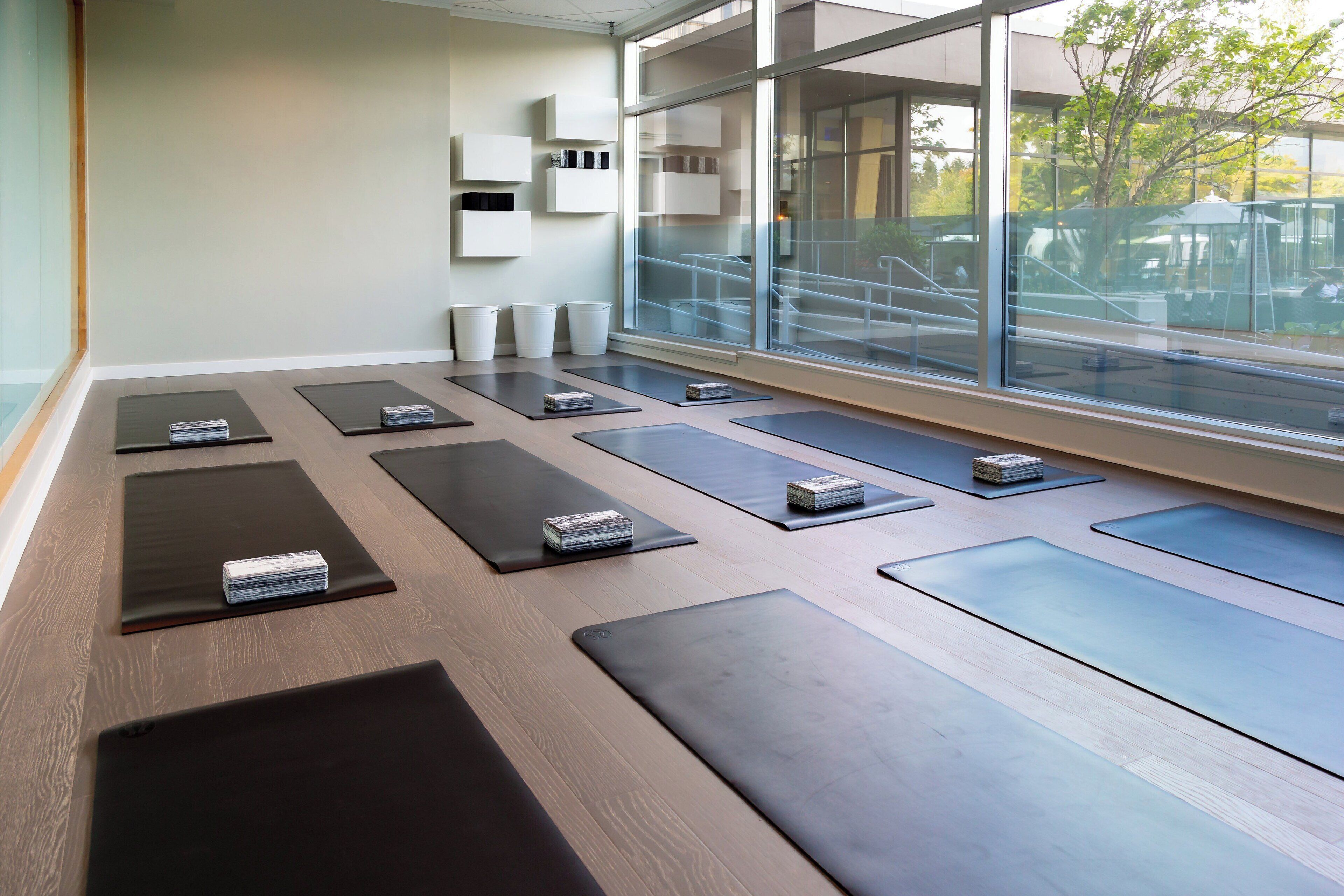 Un estudio de yoga con colchonetas dispuestas en filas, un ventanal con vistas a una terraza al aire libre, y un interior limpio y moderno.