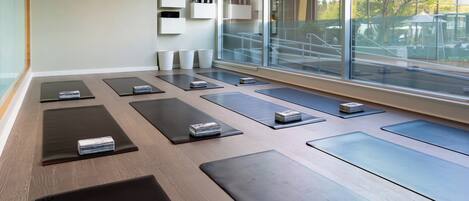 Un estudio de yoga con colchonetas dispuestas en filas, un ventanal con vistas a una terraza al aire libre, y un interior limpio y moderno.