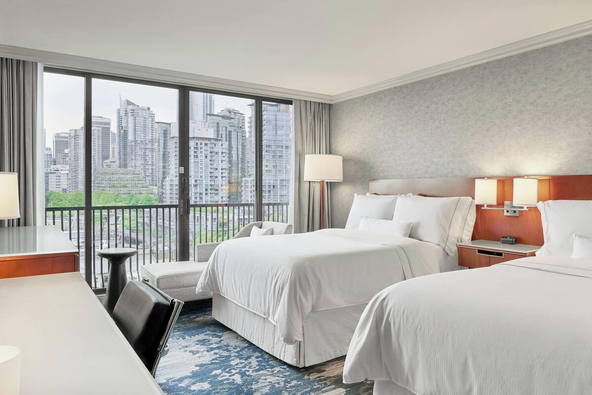 A hotel room with two beds, a desk, and a view of city buildings.