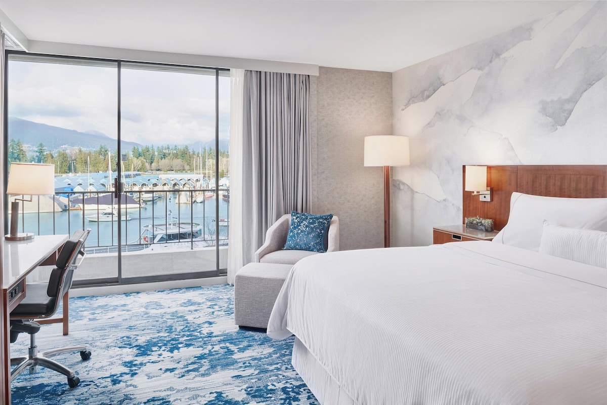 A hotel room with a large bed, a desk, a chair, a lamp, and a view of a marina.