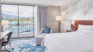 A hotel room with a large bed, a desk, a chair, a lamp, and a view of a marina.