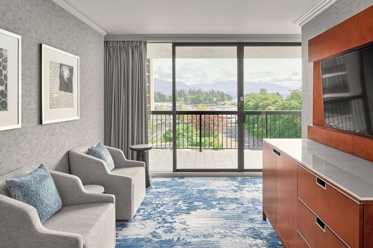 A modern living room with a grey sofa, a TV, and a large window with a view of greenery and mountains.