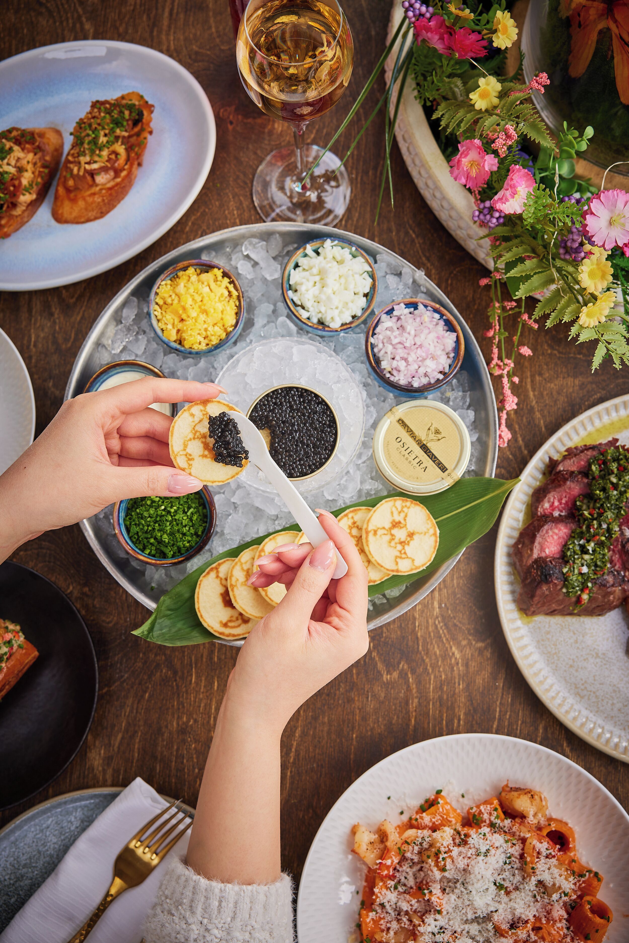 Una mesa de comedor con diferentes alimentos, condimentos, una copa de vino y un arreglo floral decorativo.