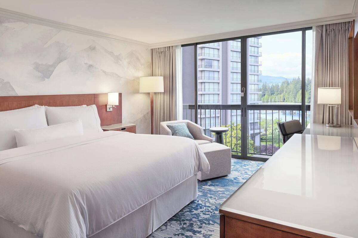 A hotel room with a large bed, a desk, a chair, and a view of a cityscape through large windows.