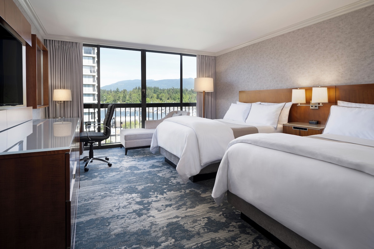 A hotel room with two beds, a desk, a chair, and a large window with a view.