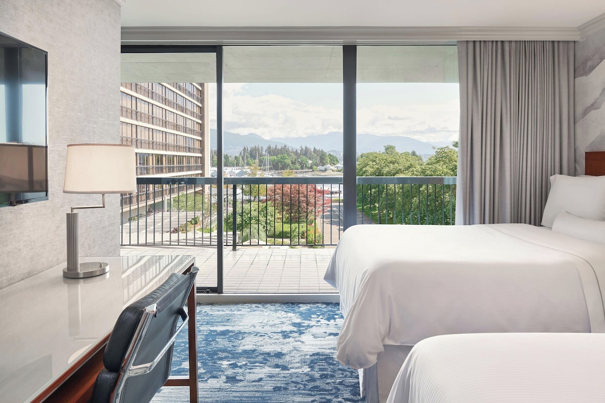 A hotel room with a large bed, a desk, a chair, a TV, and a balcony with a view of buildings and trees.