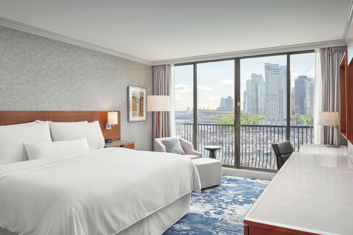 A hotel room with a large bed, a desk, a chair, a small table, and a view of the city through large windows.