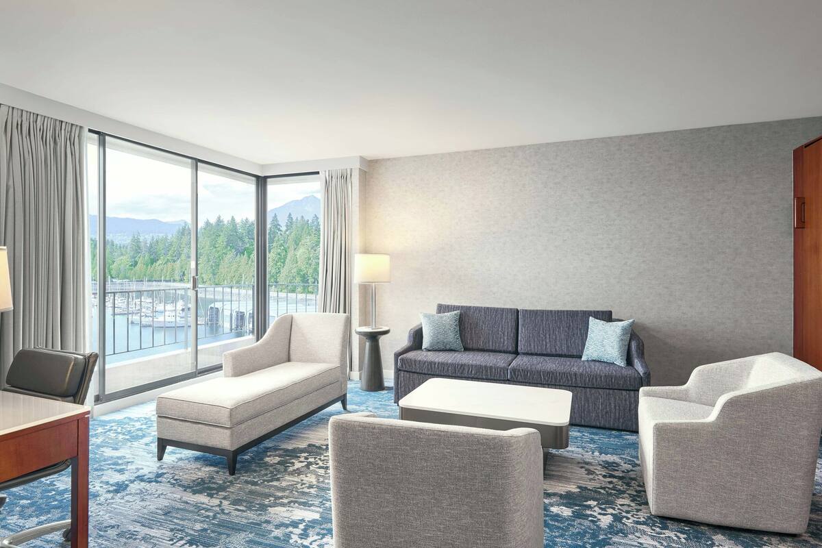 A hotel room with a large window, a sofa, a chair, a small table, and a lamp.