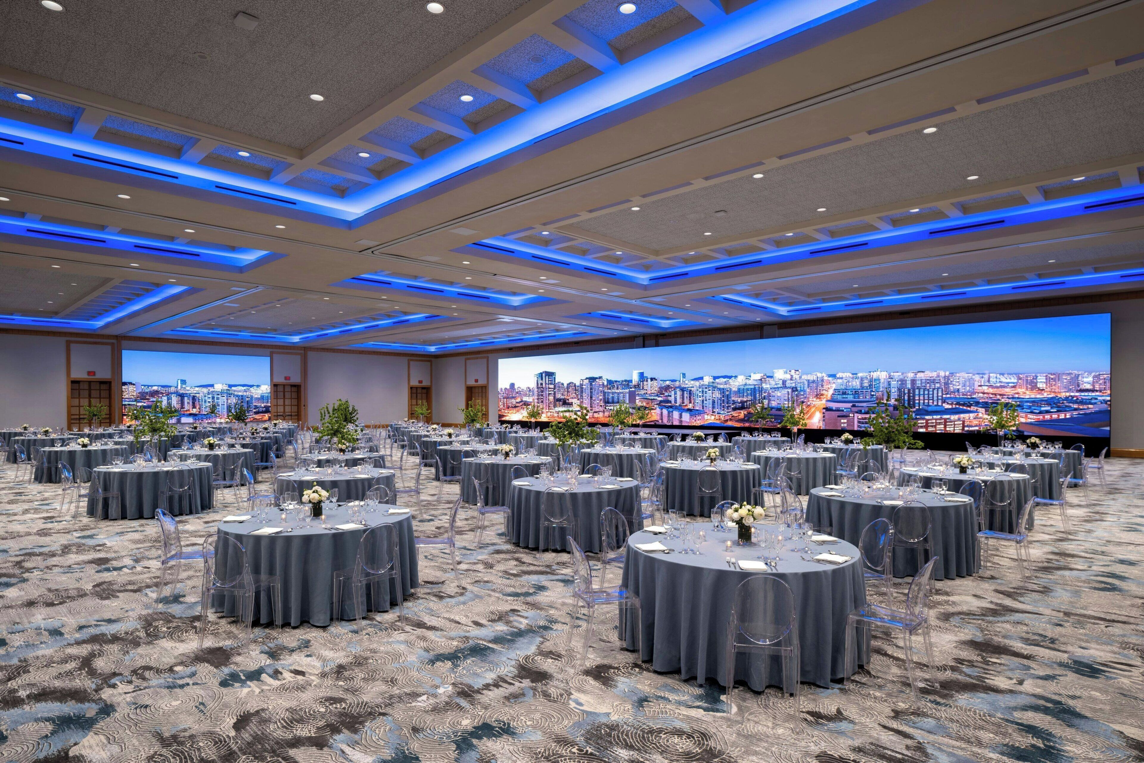 Un amplio salón de conferencias con varias mesas redondas, sillas transparentes y vistas de la ciudad en una pantalla grande.