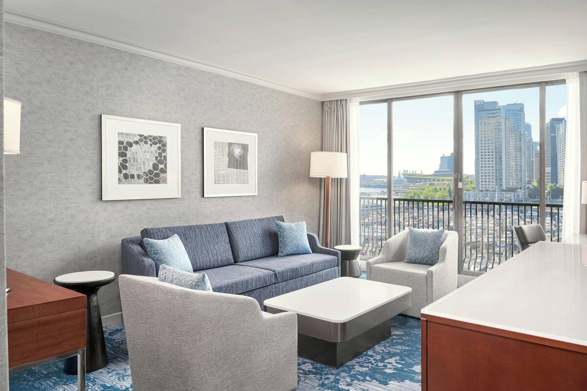 A modern hotel room with a grey sofa, a grey armchair, a white coffee table, and a desk. There are two framed pictures on the wall, a lamp, and a view of the city through large windows.