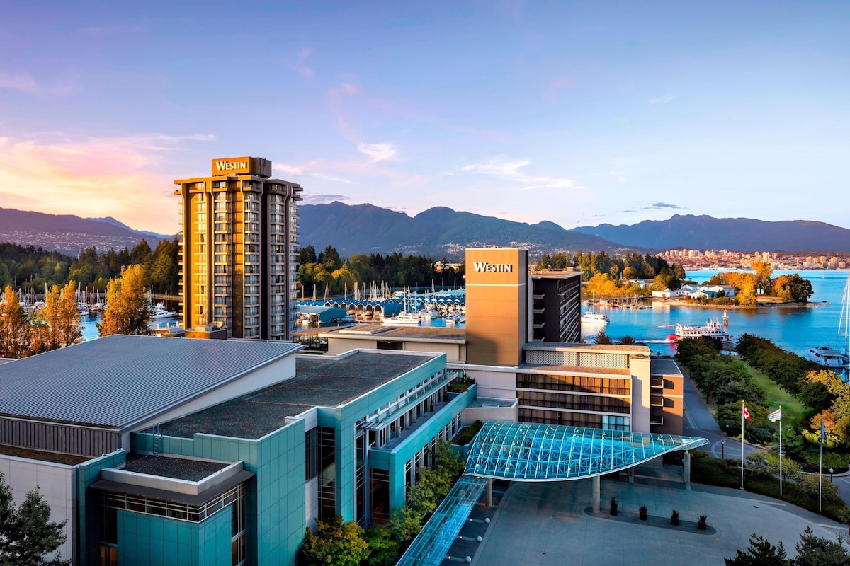 A Westin hotel with a modern architectural design, featuring a prominent building with a sign, a glass entrance, and a view of a marina and mountains in the background.