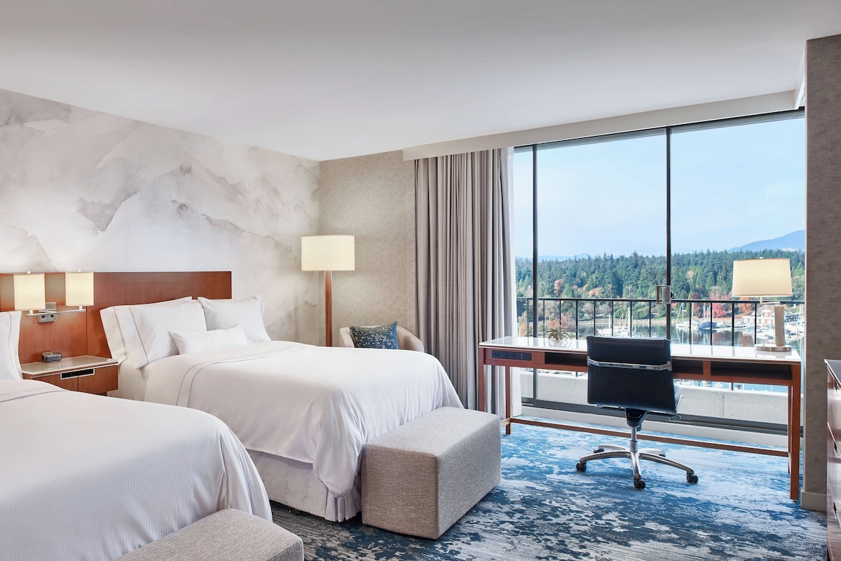A hotel room with two beds, a desk, a chair, and a large window offering a view of a forest and water.