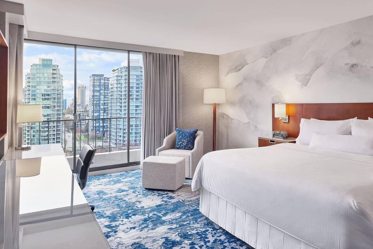 A hotel room with a large bed, a chair, a desk, and a view of tall buildings through large windows.