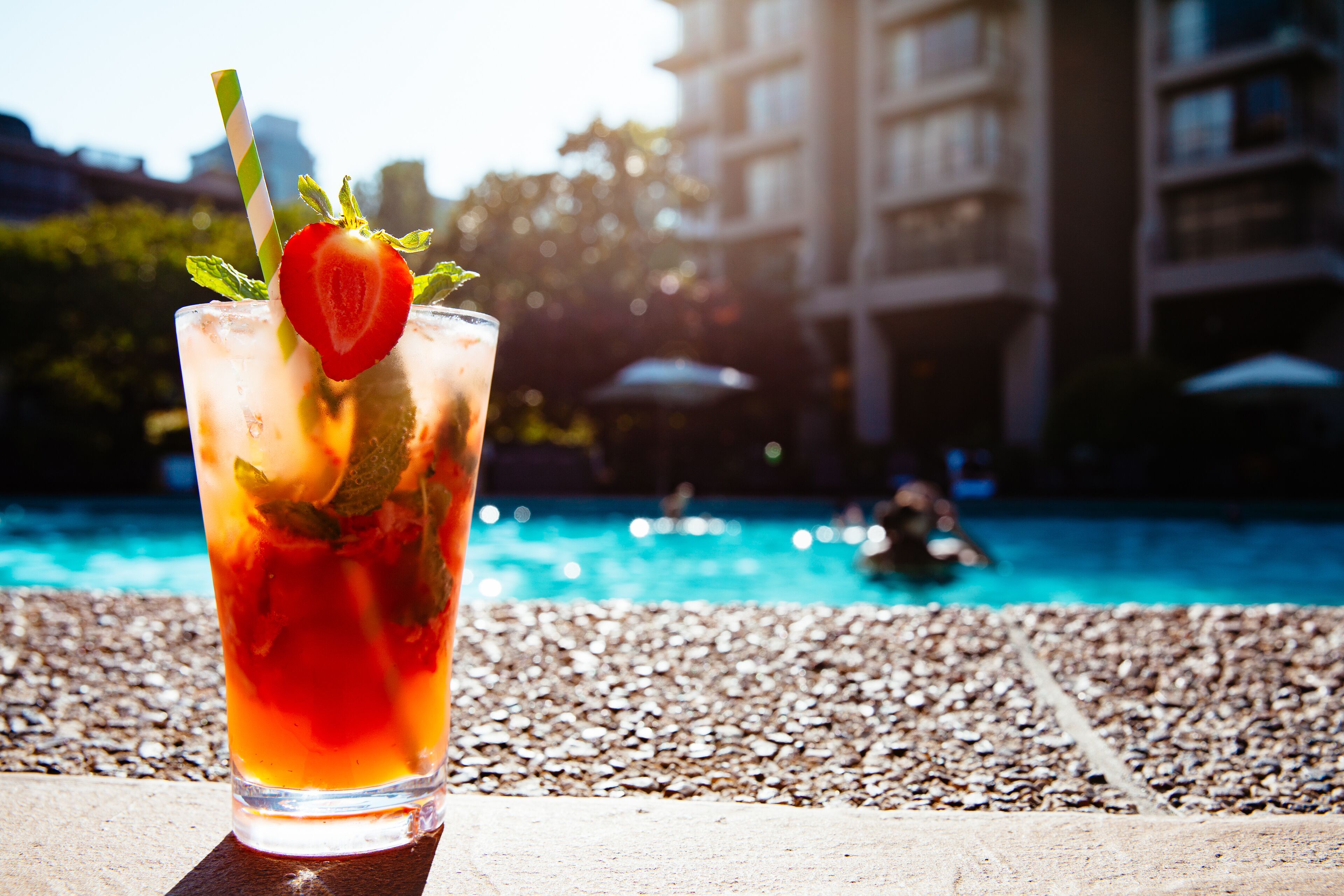 Un vaso de bebida helada adornado con fresa y menta, sobre una superficie pedregosa cerca de una piscina con un edificio al fondo.
