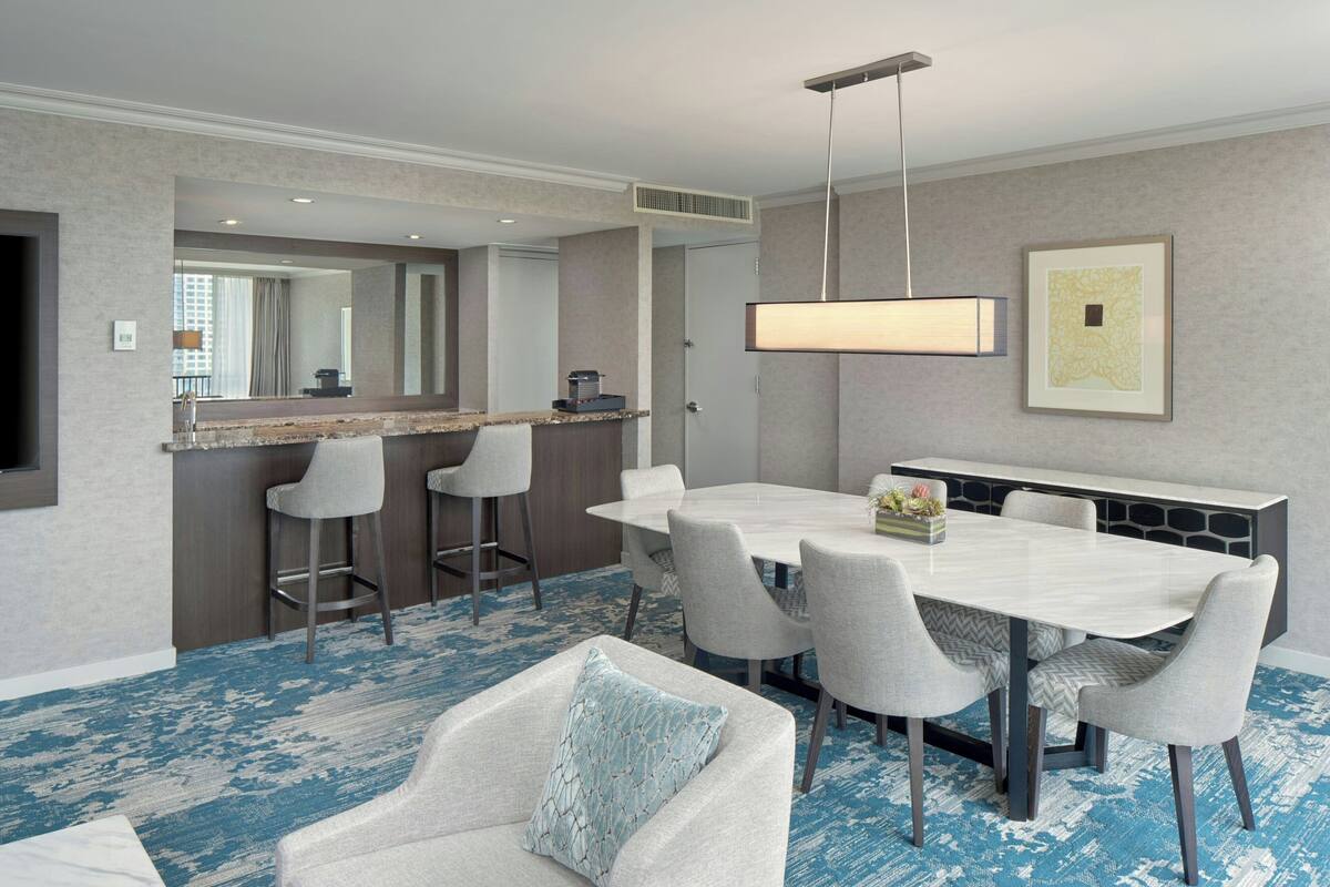 A hotel room with a dining table, bar stools, a sofa, a large mirror, and a hanging light fixture.