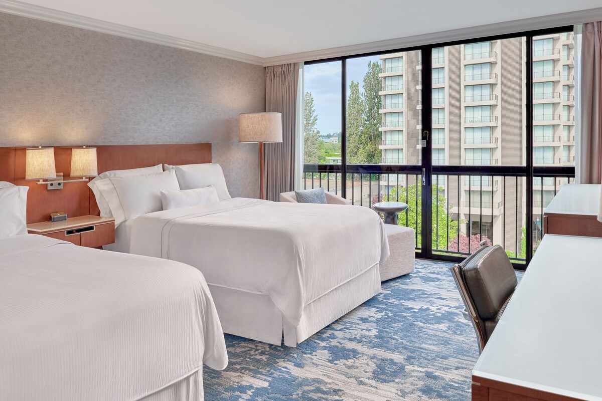 A hotel room with two beds, a desk, and a balcony with a view of high-rise buildings.