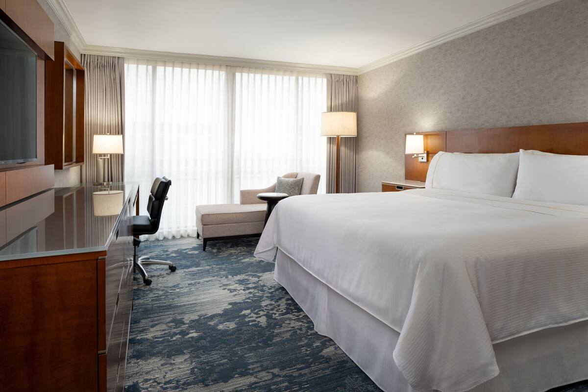 A hotel room with a large bed, a desk with a chair, a TV, and a window with curtains.
