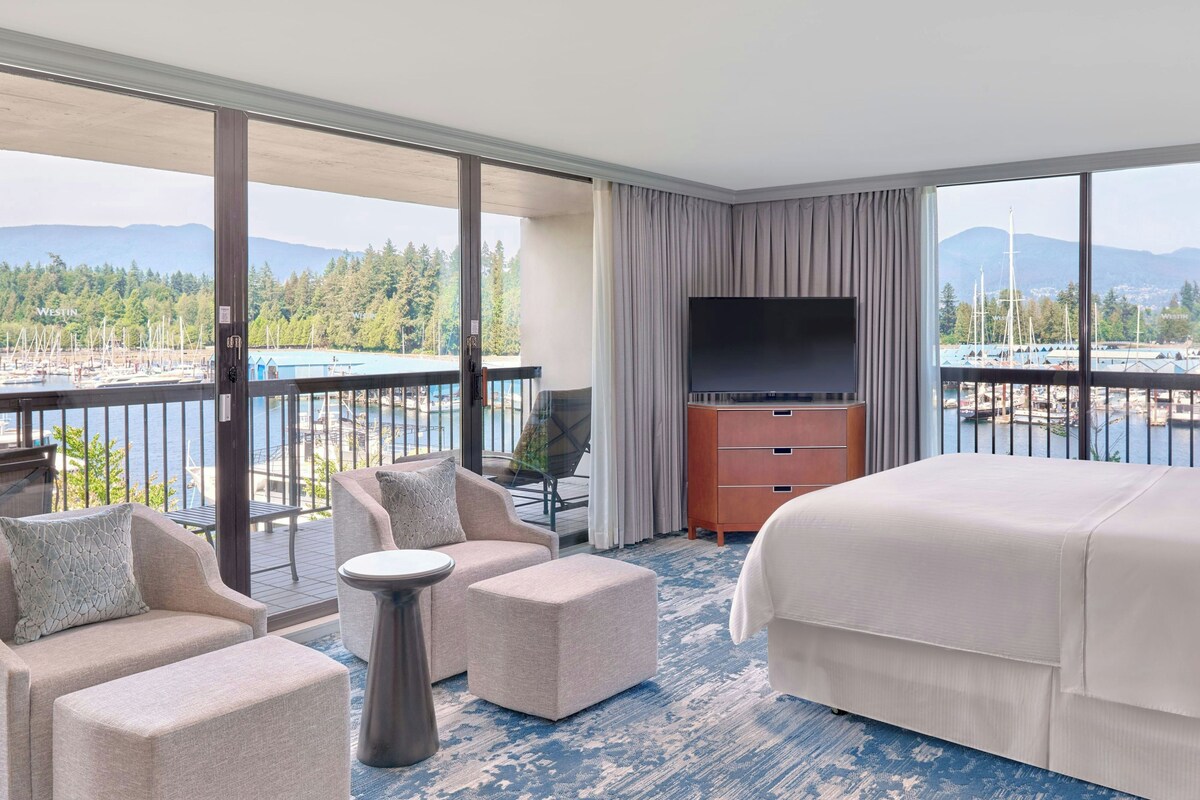Quarto de hotel com uma cama grande, televisão, área de estar com cadeiras e uma mesa, e varanda com vista para um porto de iates e montanhas.