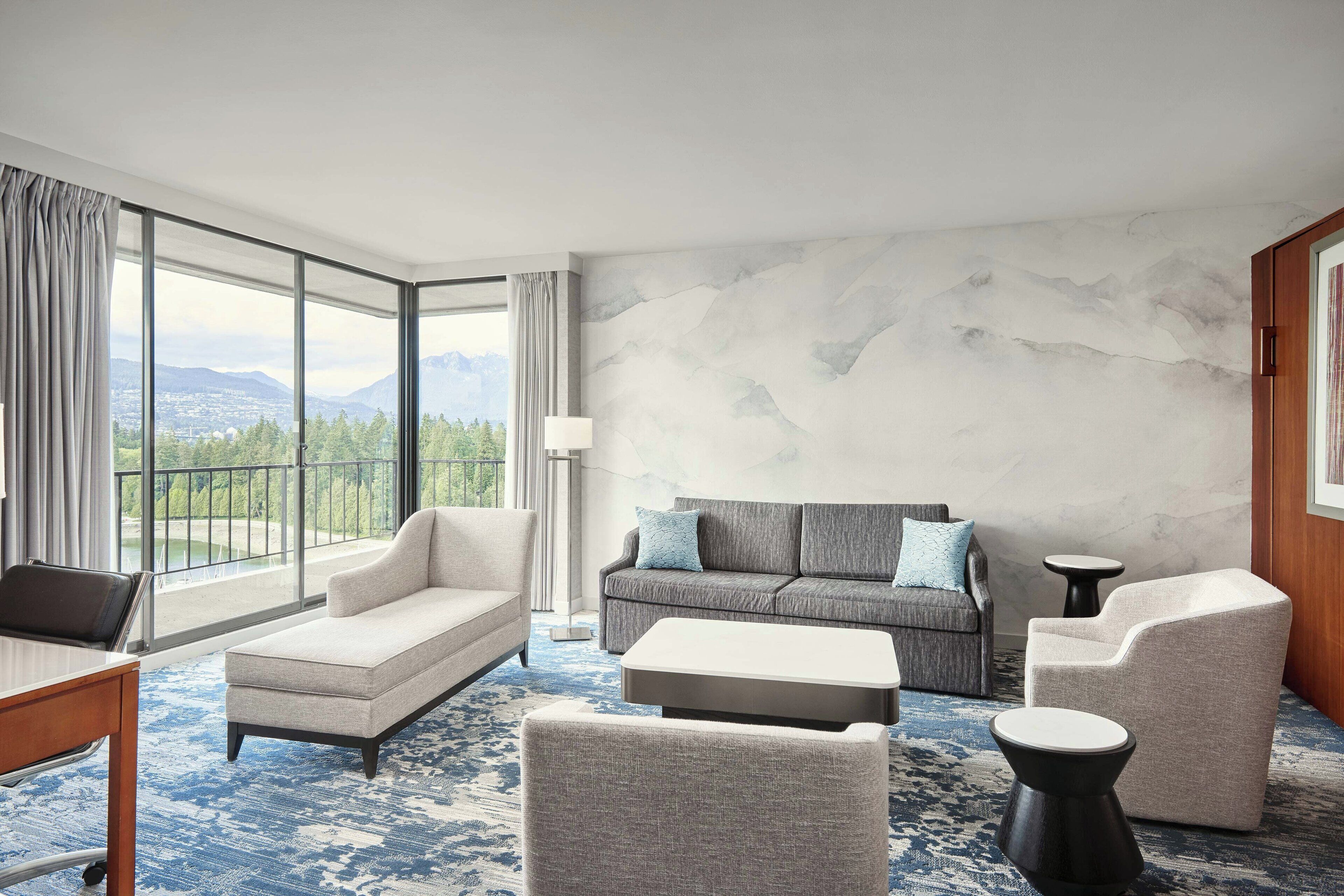 A modern hotel room with a large window offering a mountain view, a sofa, a small table, and a chair.