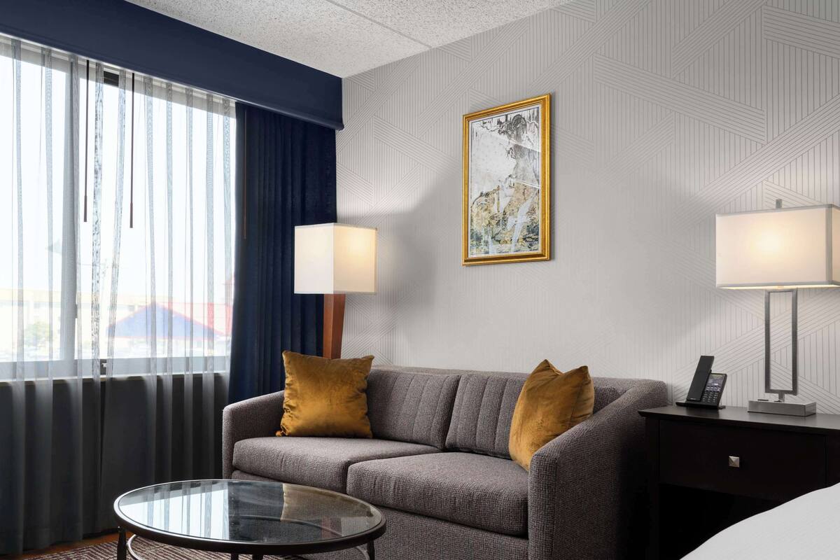 A hotel room with a gray sofa, two pillows, a glass coffee table, a bedside lamp, a wall-mounted telephone, and a framed picture.