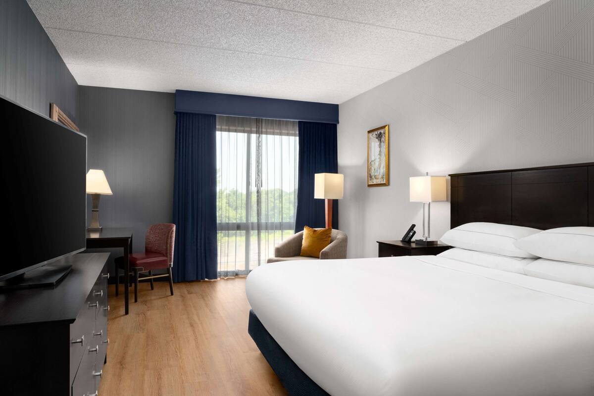 A hotel room with a large bed, a desk with a TV, two chairs, a nightstand with a lamp, and a window with curtains.