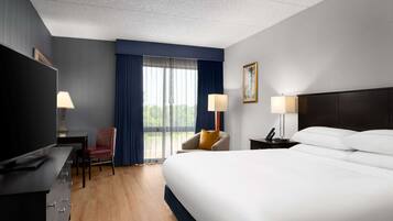 A hotel room with a large bed, a desk with a TV, two chairs, a nightstand with a lamp, and a window with curtains.