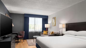 Quarto de hotel com uma cama grande, televisão, escrivaninha com luminária, cadeira e janela com cortinas.