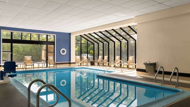 Una piscina interior con paredes de vidrio transparente, zona de piscina azul y áreas de estar iluminadas por el sol.