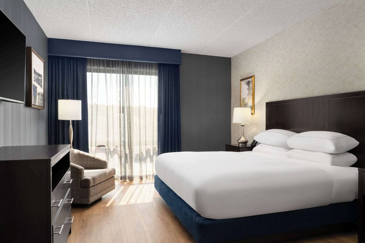 A hotel room with a large bed, a desk, a chair, a lamp, and a view of the outdoors through a window with curtains.