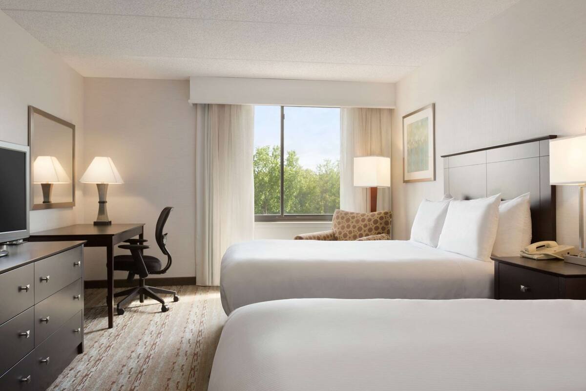 A hotel room with two beds, a desk, a chair, a TV, and a window with a view of trees.