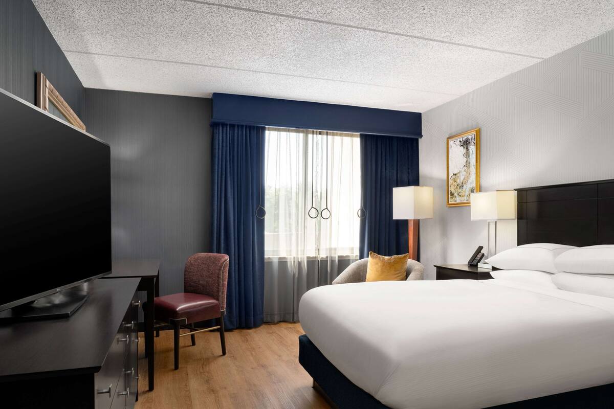 A hotel room with a large bed, a desk with a flat-screen TV, a chair, a nightstand with a lamp, and a window with curtains.