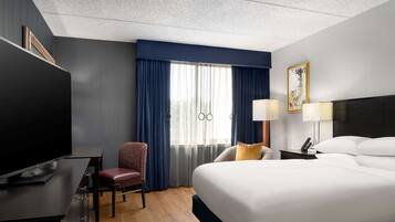 A hotel room with a large bed, a desk with a flat-screen TV, a chair, a nightstand with a lamp, and a window with curtains.