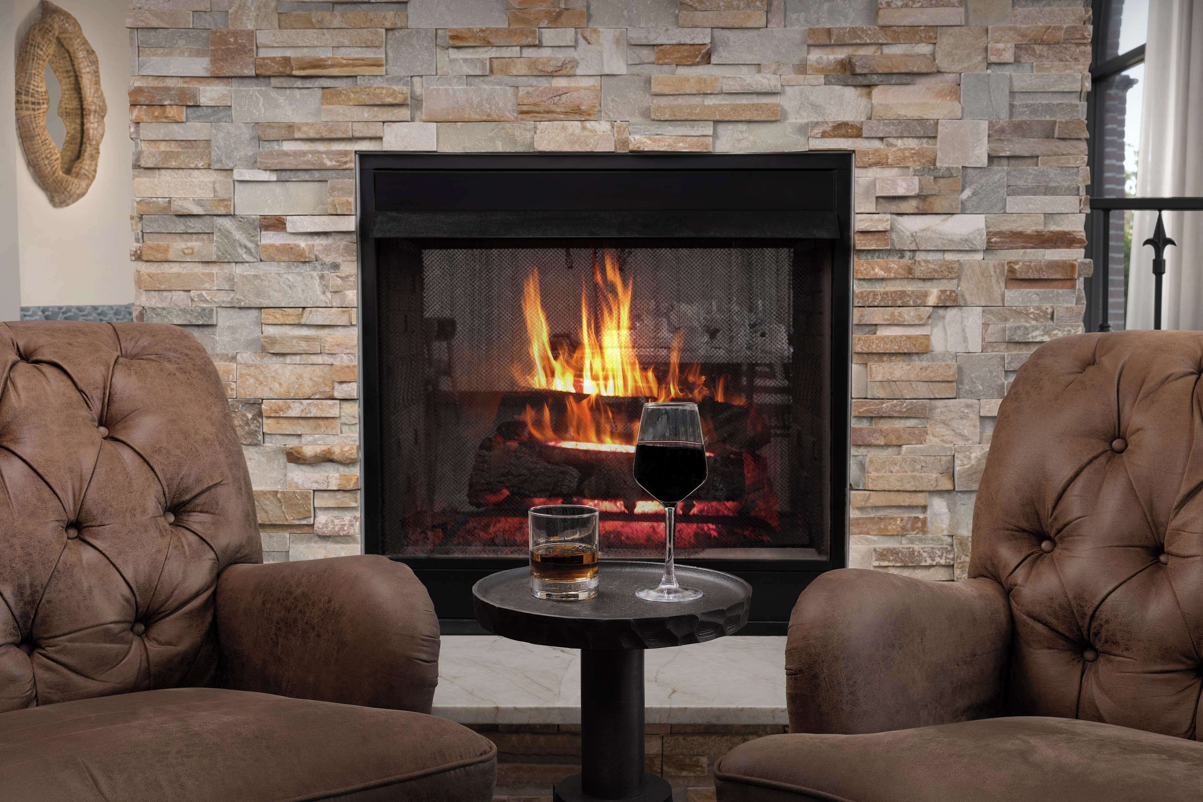 A fireplace with a stone facade, two leather armchairs, and a table with a glass and a bottle.