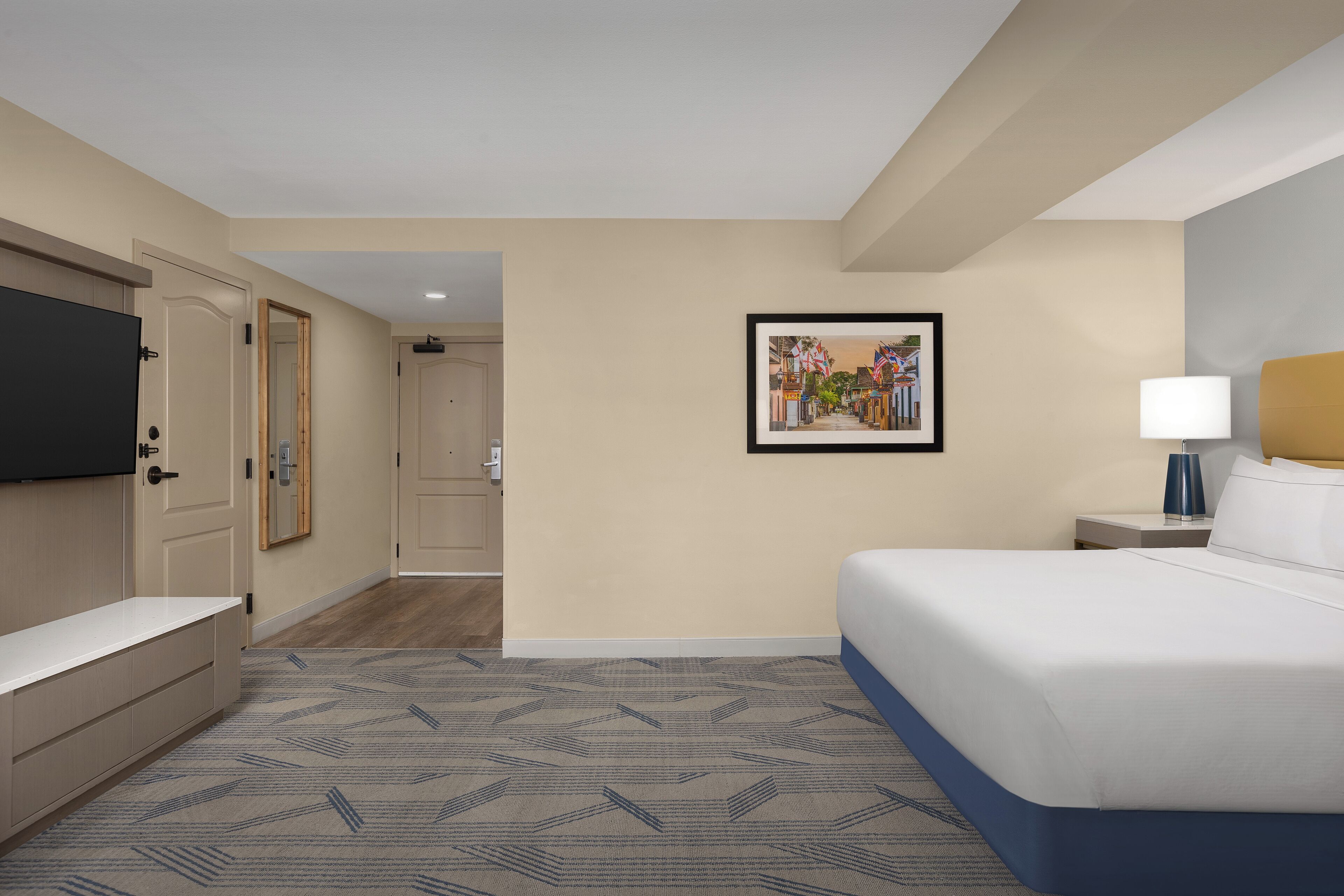 A hotel room with a bed, bedside table, lamp, and a framed picture on the wall.