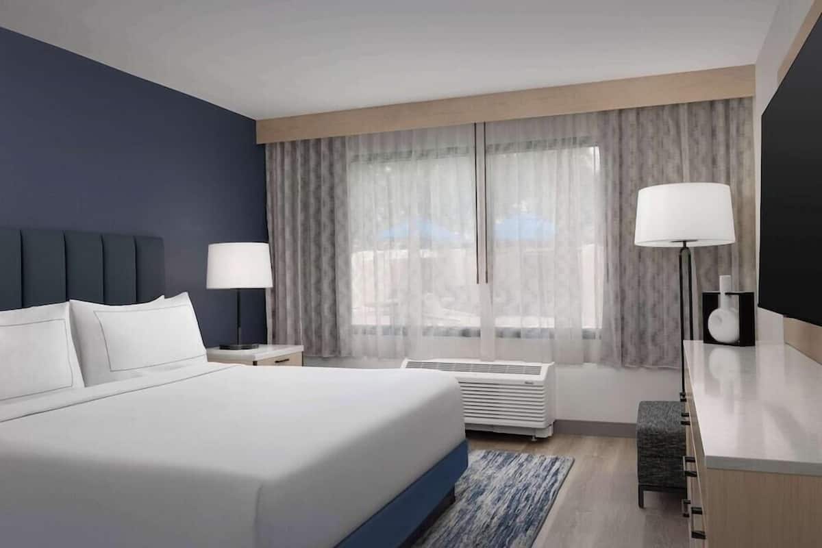 A modern hotel room with a large bed, a desk, a chair, a lamp, and a window with curtains.