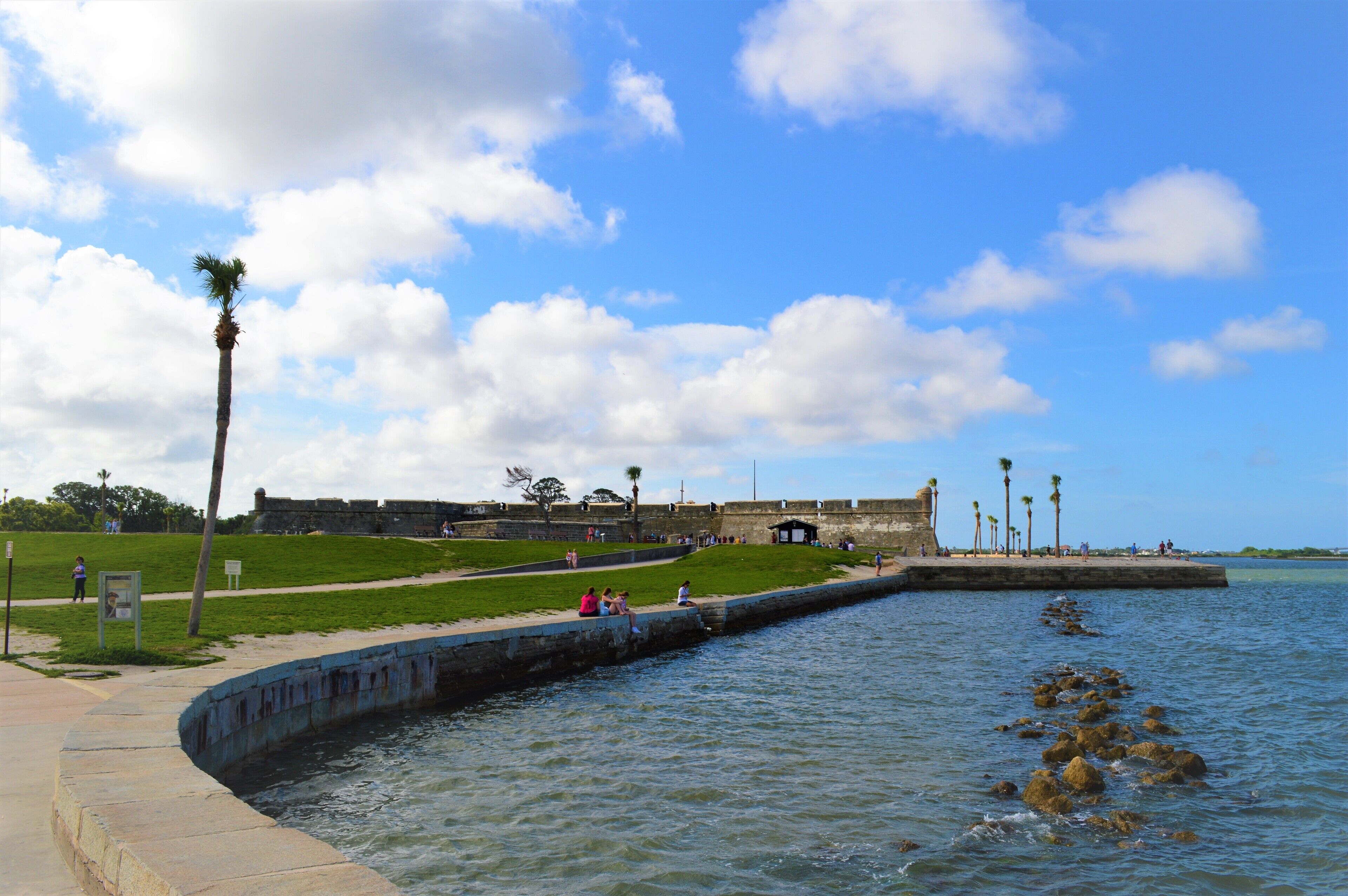 Una fortificació costanera amb una zona verda, palmeres i una paret de pedra a la vora del mar.