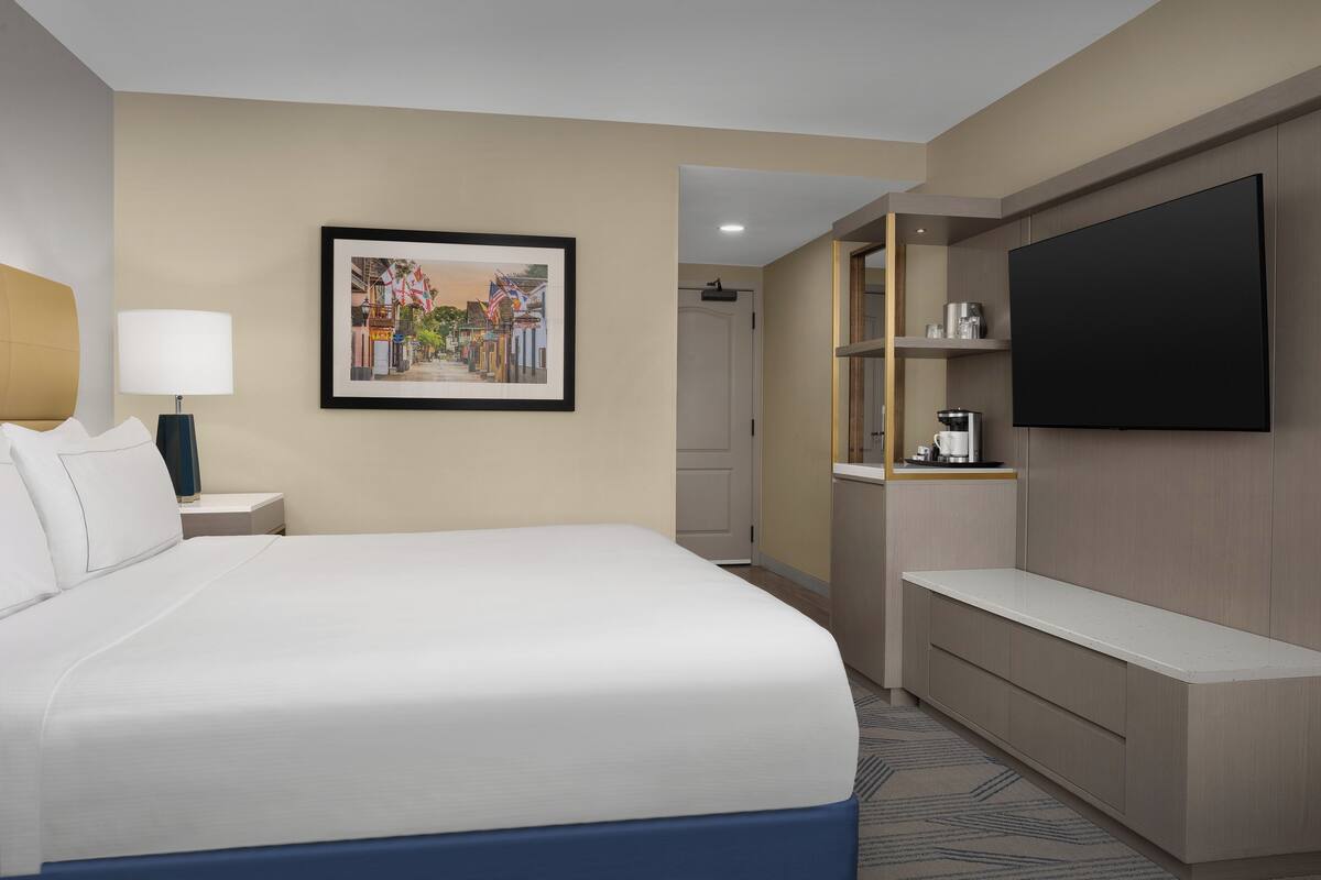 A hotel room with a large bed, a TV mounted on the wall, a coffee machine, and a framed picture on the wall.