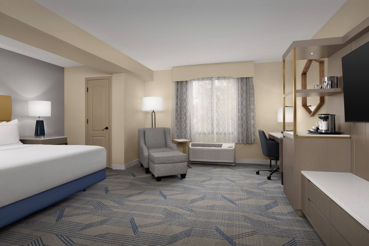 A hotel room with a bed, a desk, a chair, a TV, and a window with curtains.