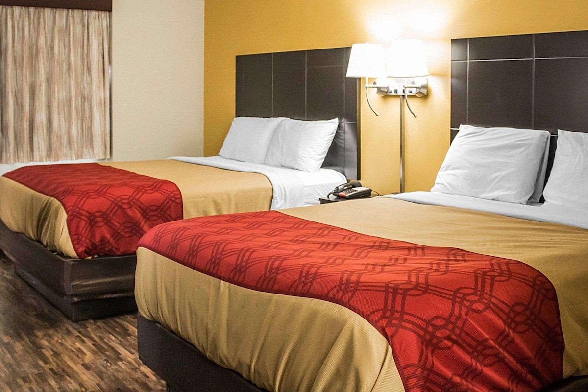 Hotellihuone, jossa on kaksi sänkyä, joissa kummassakin on punavalkoinen ja beigenvärinen peitto, yöpöytä lampun kanssa sekä ikkuna, jossa on verhot.
