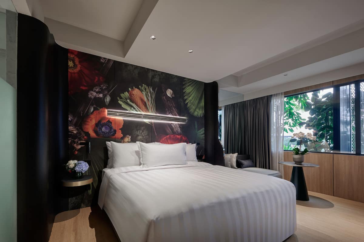 Une chambre d’hôtel moderne avec un grand lit, une table de chevet ornée d’un vase de fleurs, une petite table ronde et une vue sur la verdure par la fenêtre.