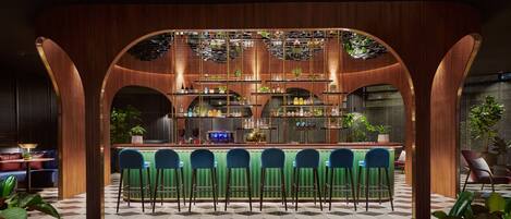 Zona de bar con un fondo de madera, varias estanterías con botellas y taburetes de bar verdes.
