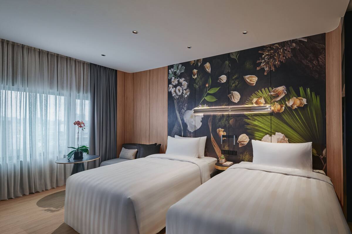 Une chambre d’hôtel avec deux lits, une grande fenêtre avec des rideaux légers, une table ronde ornée d’un pot de fleurs et une fresque murale représentant des fleurs et des feuilles.