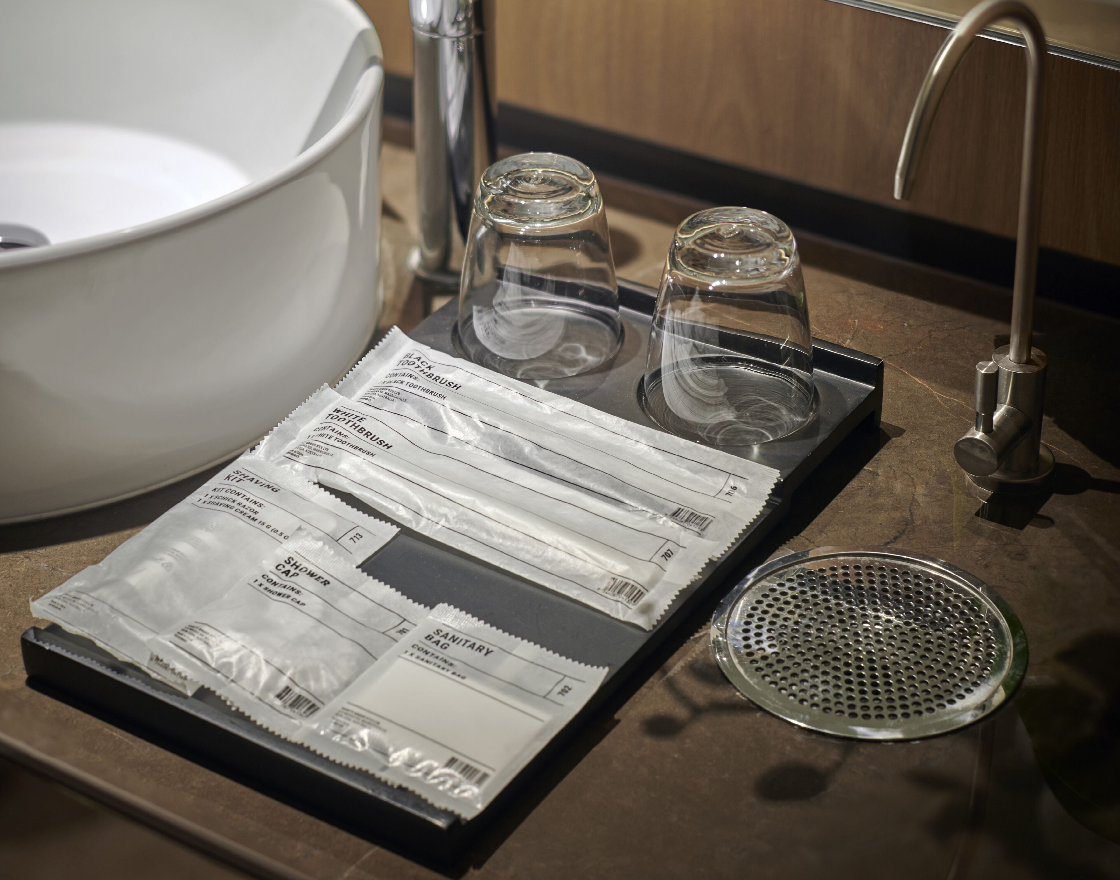 A bathroom sink with a white basin, two clear glass cups, a metal strainer, and a package of sanitary bags.