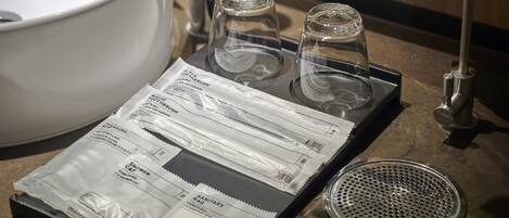 A bathroom sink with a white basin, two clear glass cups, a metal strainer, and a package of sanitary bags.