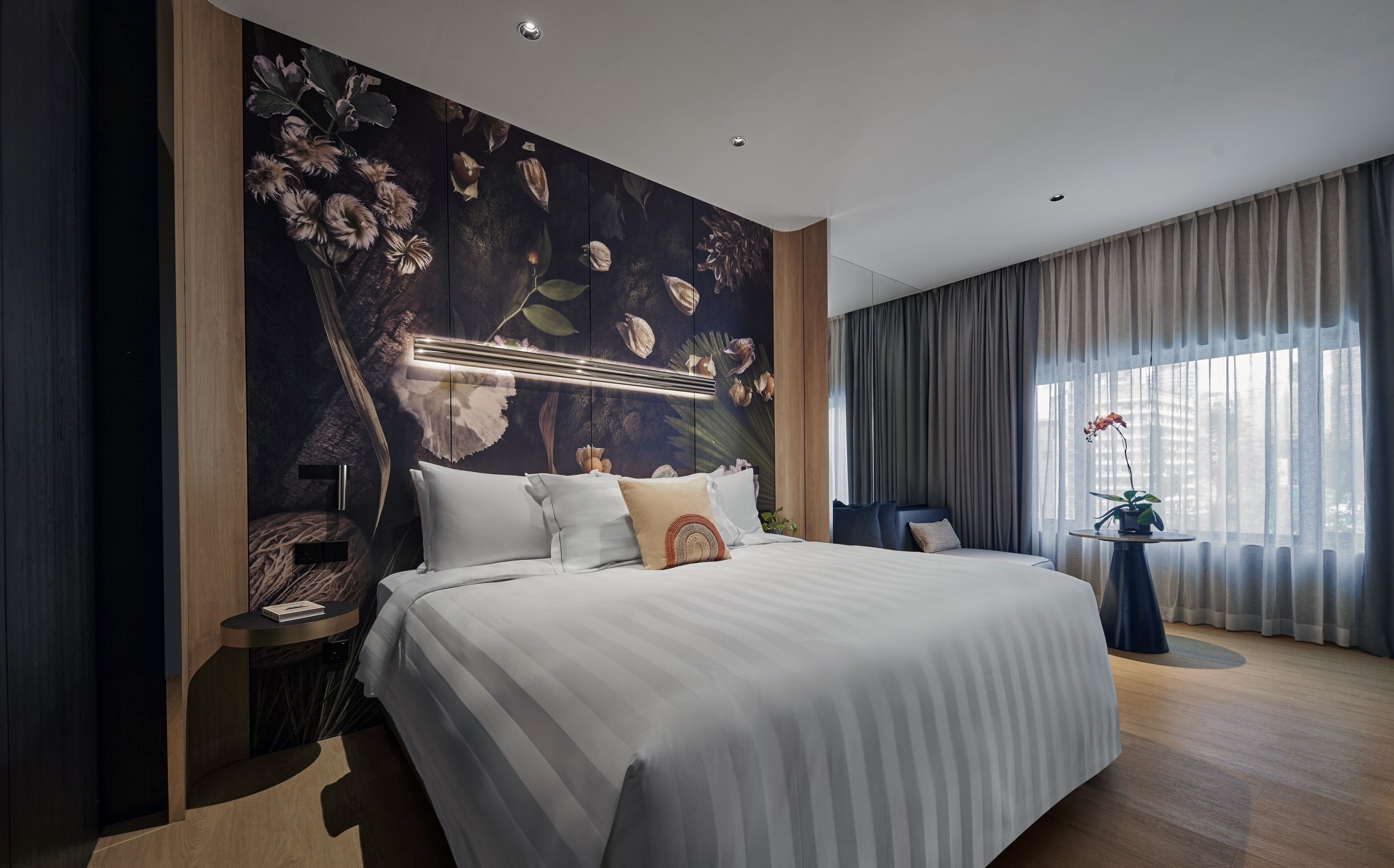 A modern hotel room with a large bed, a nightstand, a small table with a plant, and a view of the city through the window.