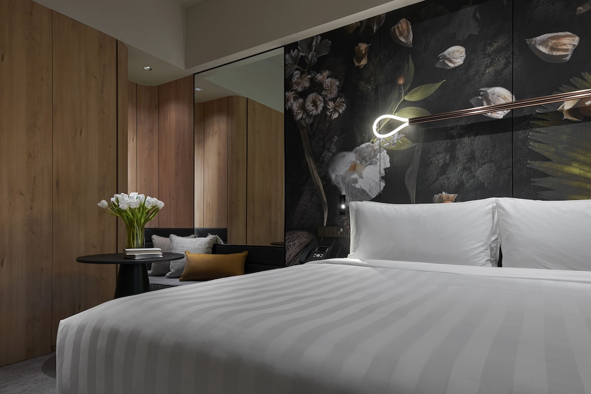A modern bedroom with a large bed, a bedside table with a vase of flowers, and a wall-mounted mirror.