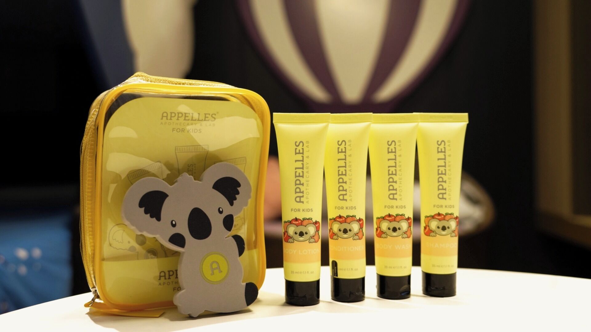 A yellow toiletry bag with a bear cutout and four tubes of Apelles body care products.