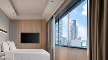 A modern hotel room with a large bed, a flat-screen TV mounted on the wall, and a city view through large windows.