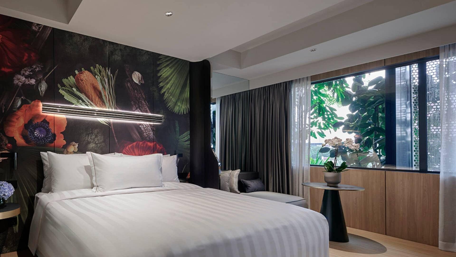 A modern hotel room with a large bed, a wall mural, a window with a view of greenery, and a small table with a plant.